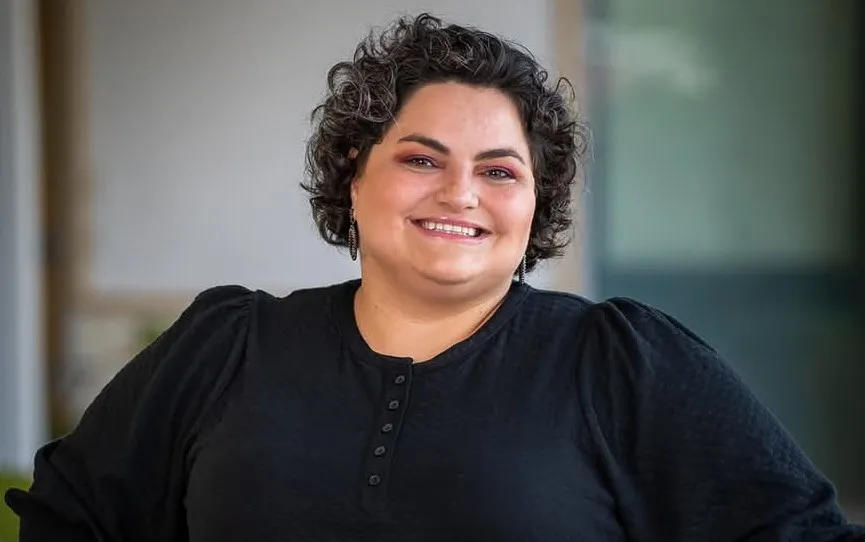 The image features a smiling woman with curly dark hair, wearing a black, buttoned shirt and dangling earrings.