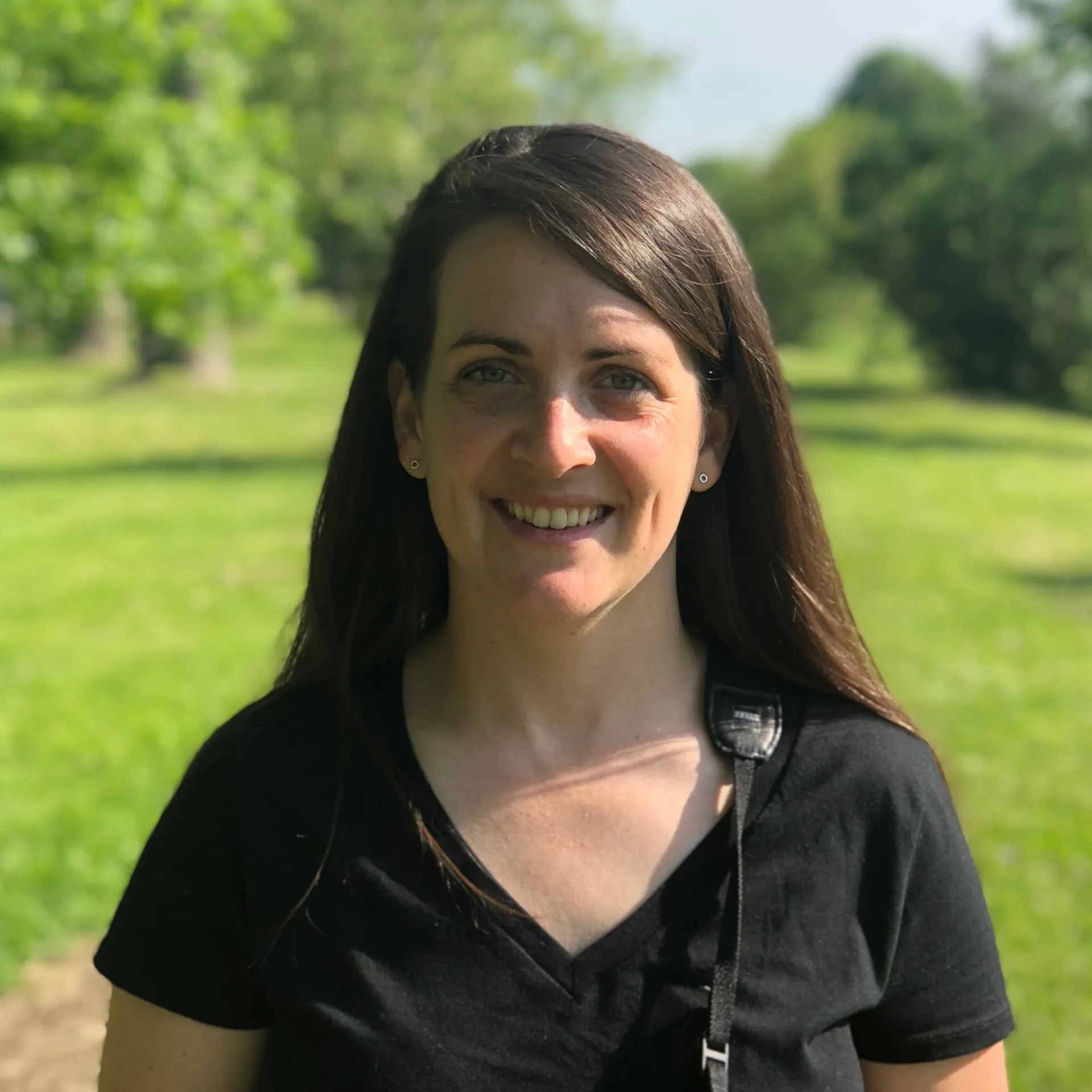 A headshot photo of a brown-haired woman standing in a grassy field