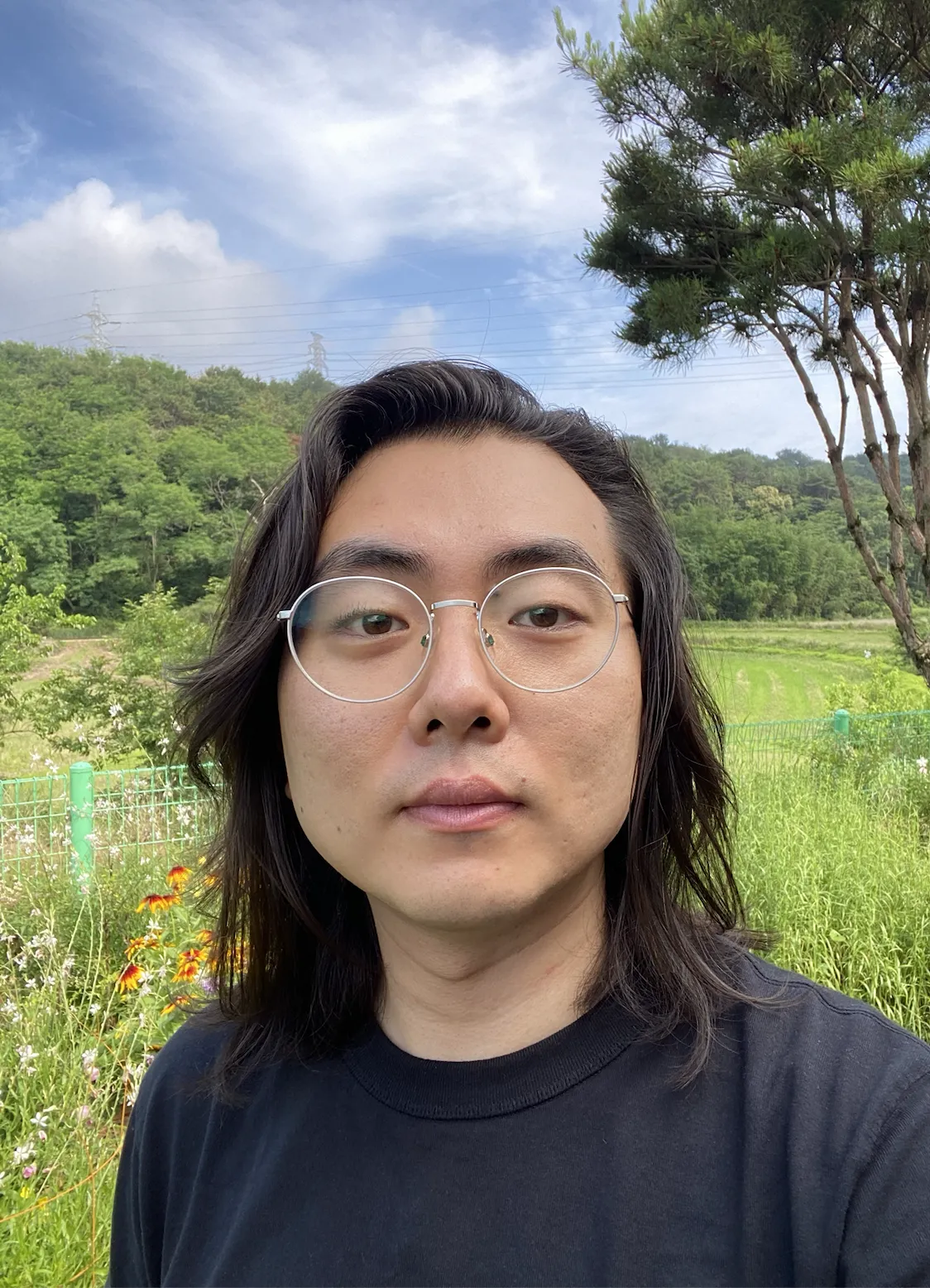 Headshot of a man against a backdrop consisting of trees, flowers, and a blue sky with clouds.