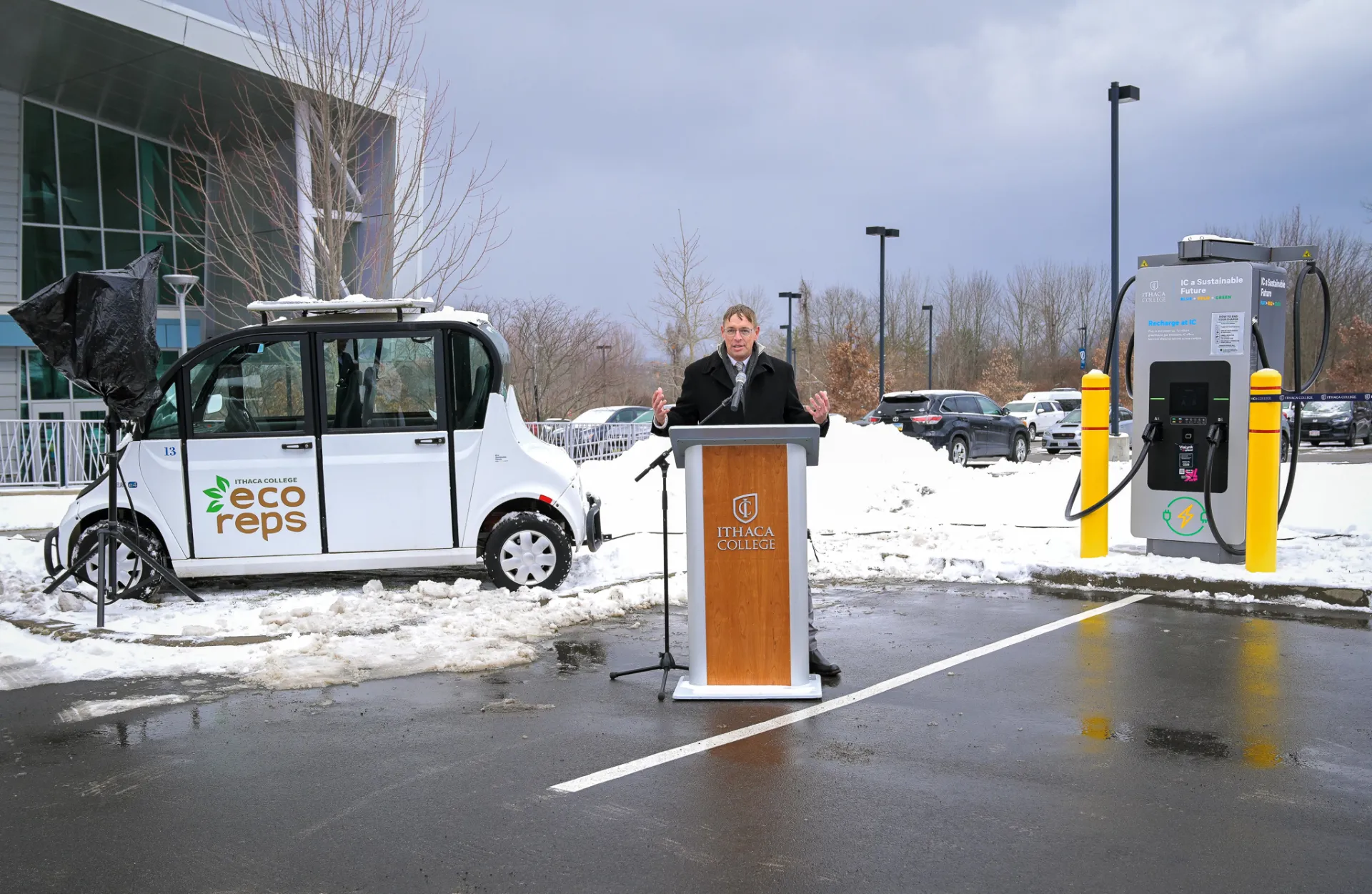 A man stands at a podium on a snowy day between an EV and an EV charger.