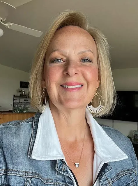 Photo of head and shoulders of a woman looking directly in camera. Blond hair to shoulder length, blue eyes. Wear a white collared shirt with a jean jacket 