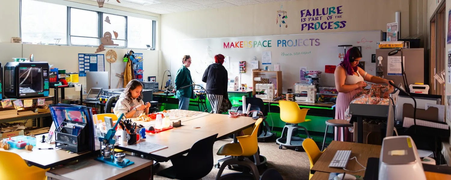 Wide shot of the Makerspace room - it's very full of stuff, including 3D printers, sewing machines, a button maker, and large worktables in the center. A students sits at the center table working on a project, behind her two people work at the embroidery machine, and on the right side of the room a student cuts fabric. There are signs on the back wall that say "Makerspace Projects" and "Failure is part of the process".
