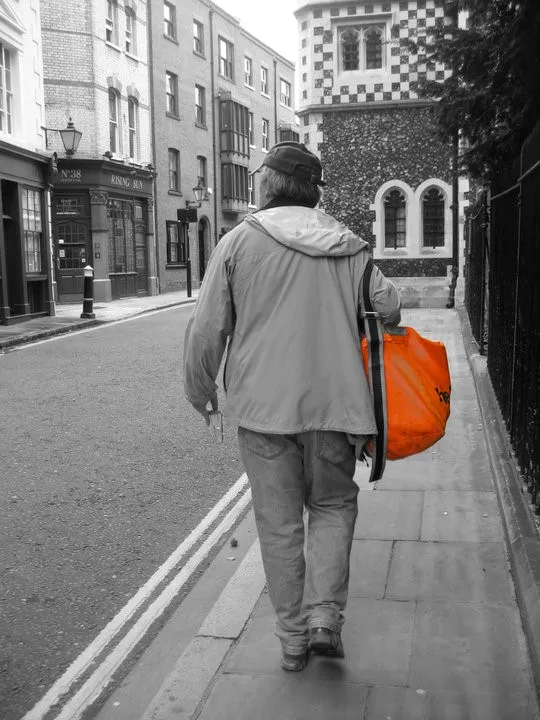 Bill Sheasgreen walking down a street in London with his back turned towards the camera. The photo is in black and white, but Bill is carrying a neon orange messenger bag on his shoulder.