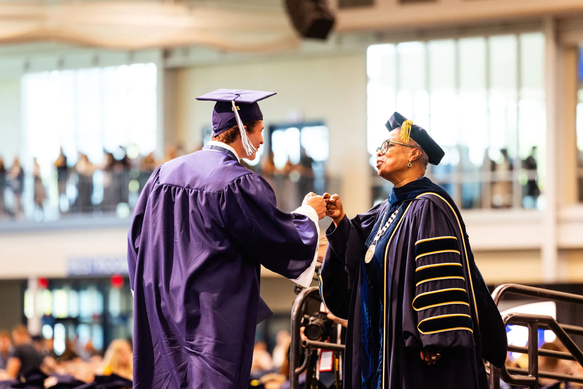 Two people in caps and gowns fist-bump at Commencement.