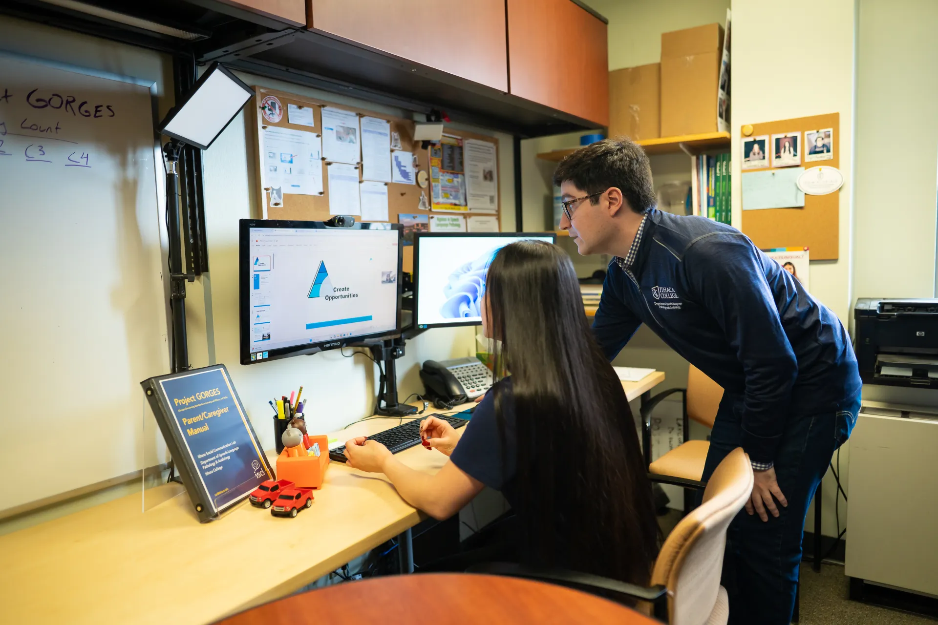 A professor and student look at a computer screen at a desk.