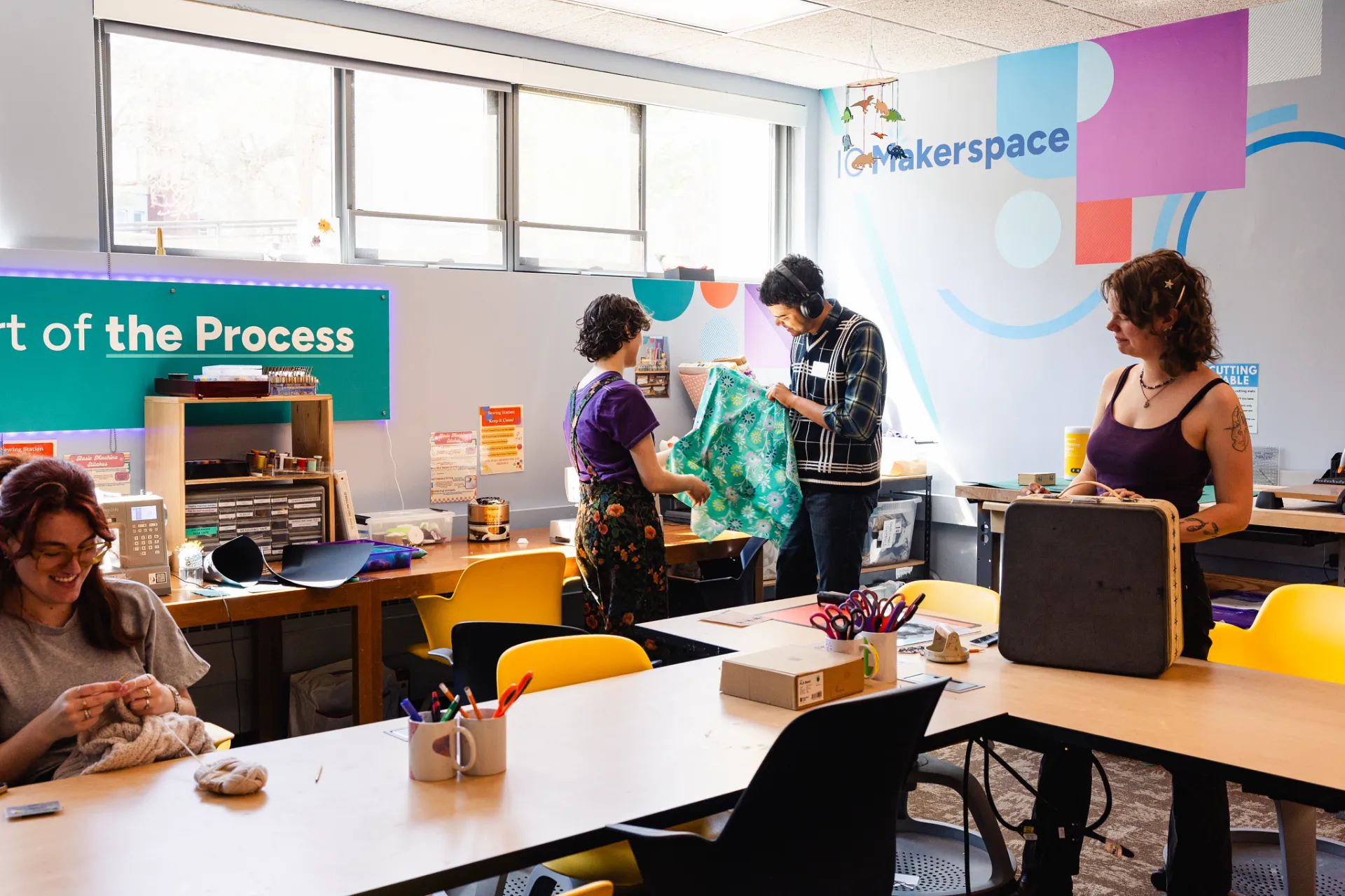 Wide shot of the Makerspace. On the left, a person is sitting at a table crocheting. In the center, two people are holding up and looking at a green piece of fabric. On the right, a person is doing printmaking. 