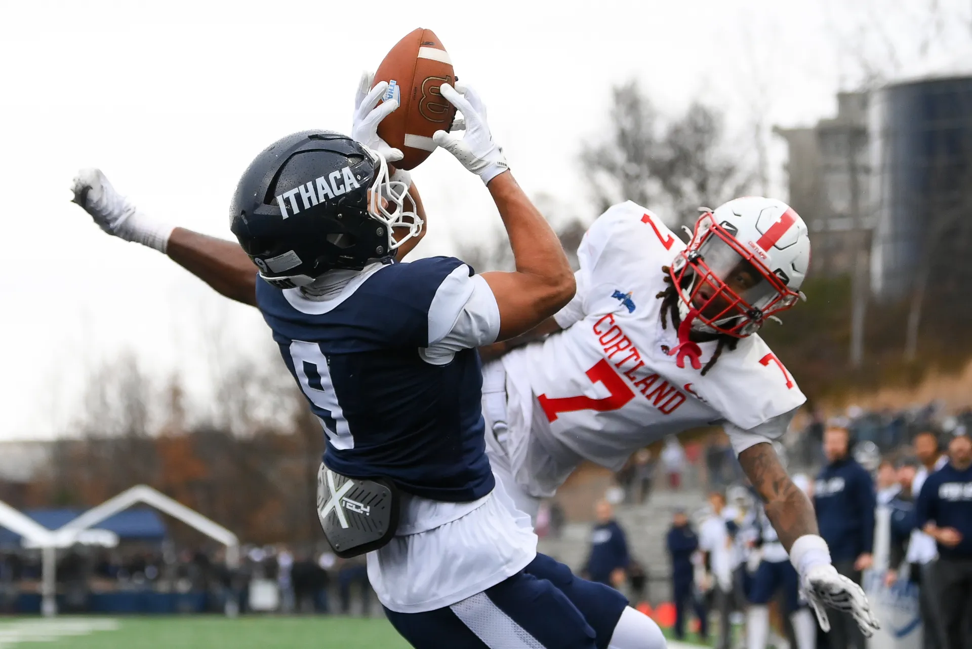 A college football player in a blue uniform catches a pass against a defender in a red uniform.