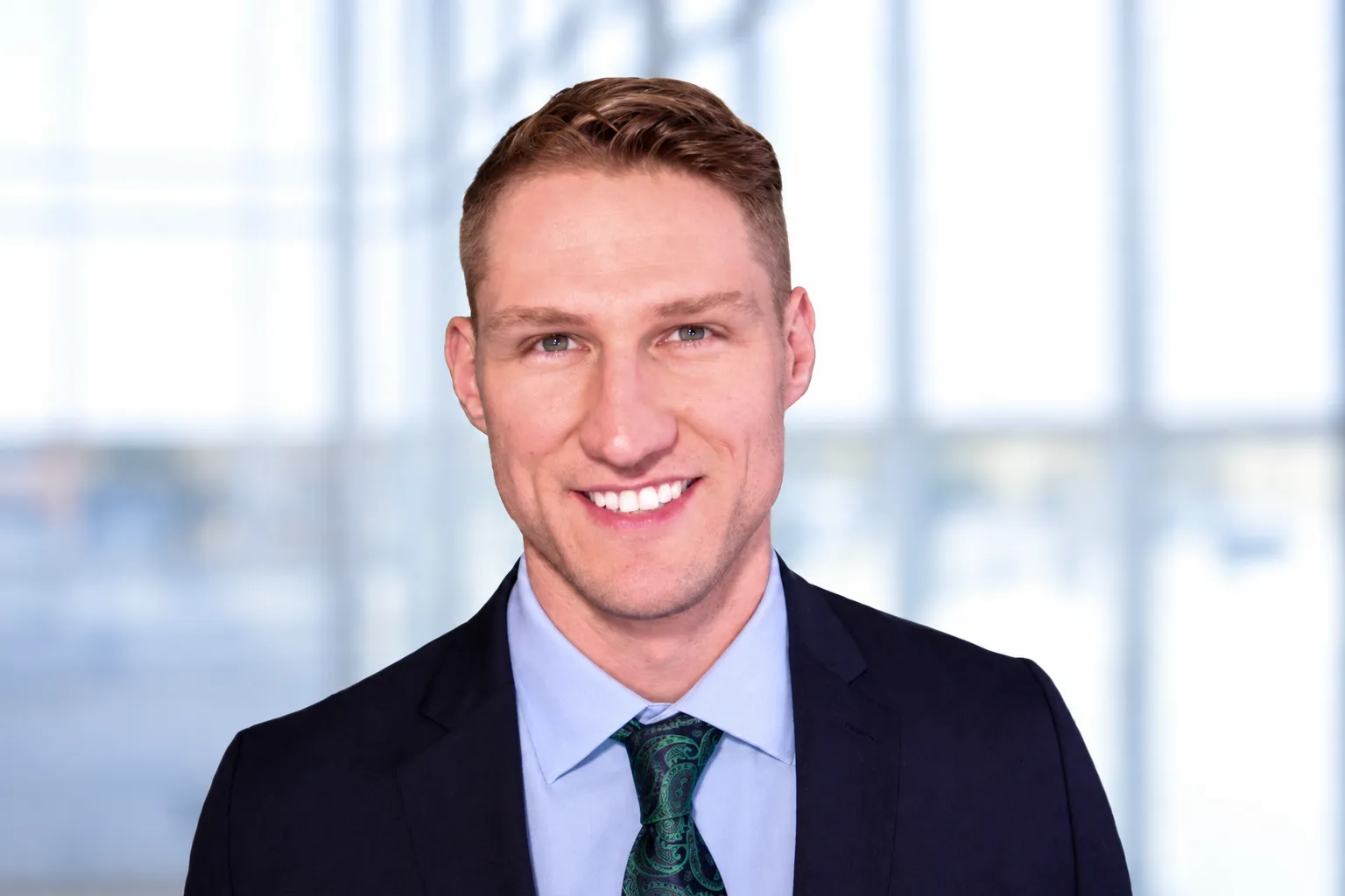 Matthew Clauhs smiling at the camera, wearing a dark suit jacket, light blue shirt, and patterned tie, with softly blurred windows in the background.