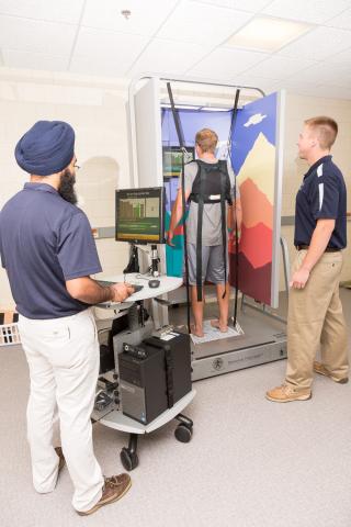 A student is in a harness attached to sensors. Another student in a blue shirt and tan pants is standing to the side observing. A faculty member is standing to the right using a computer to monitor activity.