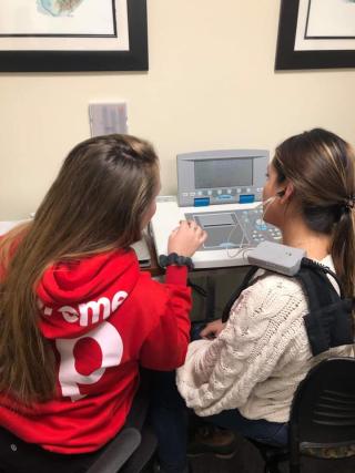 Two students have their backs to the camera. One is wearing a red shirt and the other a white shirt. They are using an audiology machine.