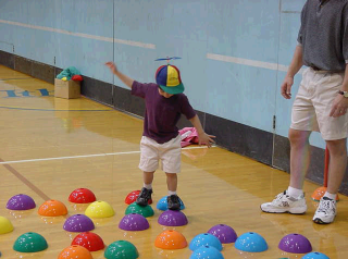 A small child is in a physical education class and is balancing on half-discs that are laid across the gymnasium floor.