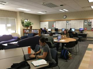 A picture of a large room with couches, work tables, and desktop computers. There are students scattered throughout the room studying.