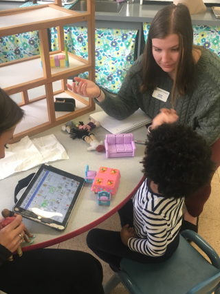 A student in a gray sweater is working with a child seated at a table. There is an ipad with information on it and the student is gesturing at the child encouraging them to communicate.