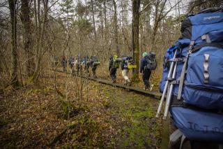 There are about 10 students wearing coats with camping backpacks on hiking through a forest.