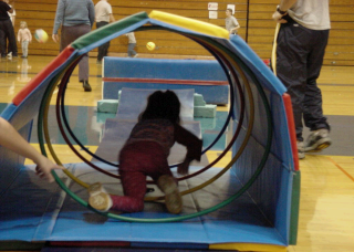 A young child is crawling through a colorful tunnel made of mats in a school gymnasium.