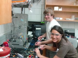two students grin at the camera while working with a tangle of wires in front of a machine