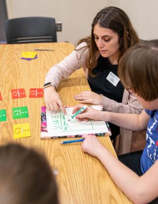 A student in a blue shirt and white sweater is seated at a table with a client heling them with a score sheet. The client is making tally marks on the score sheet related to the game they are playing.