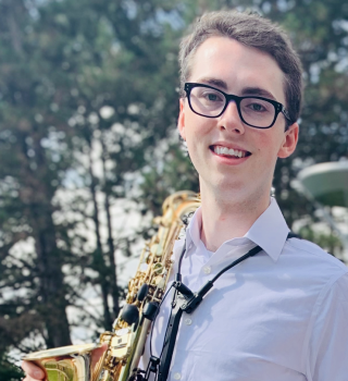 Student with alto saxophone standing outside and smiling at the camera.