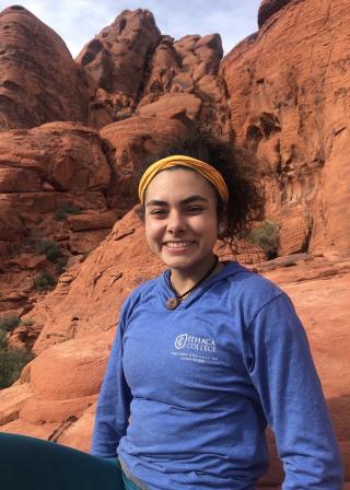 This is a photo of Angelique Atlas. Angelique is wearing a long sleeve blue shirt of Ithaca College. Angelique has a yellow hair band in the hair. Angelique is seated among a rocky landscape, out west. 