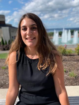 This is a photo of Chasse Guerrera. Chasse is seated in front of the Ithaca College fountains wearing a black sleeve dress.