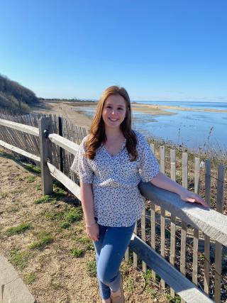 This is a photo of Danielle Frankel. Danielle is standing near a fence overlooking a lake. Danielle is wearing a flowered short-sleeve shirt and jeans.