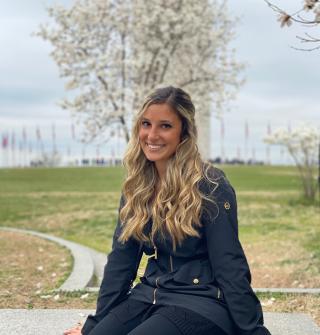 This is a photo of Danielle Pasquale. Danielle is seated outside, with green grass and a tree behind where Danielle is sitting. Danielle is wearing a black coat and pants.