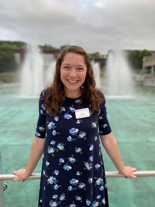 This is a photo of Elena Tansley. Elena is standing in front of the Ithaca College fountains wearing a dark blue dress with white flowers.