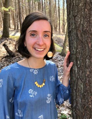 This is a photo of Elizabeth Jesch. Elizabeth is standing in the forest and is wearing a light blue shirt and white flowers.