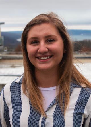 This is a photo of Emily Condellow. Emily is wearing a blue and white vertical striped shirt with a white t-shirt underneath. Emily is smiling.