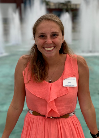 This is a photo of Haley Ashton. Haley is standing in front of the Ithaca College fountains wearing a salmon colored dress with a gold belt.
