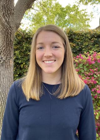 This is a photo of Hannah Thomas. Hannah is smiling at the camera with long blond hair. Hannah is wearing a long sleeve blue shirt. Hannah is standing outdoors, in front of a tree.
