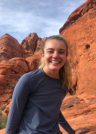 This is a photo of Jillian Boylan, standing in a rocky landscape likely out west. Jillian is wearing a long sleeve blue shirt.