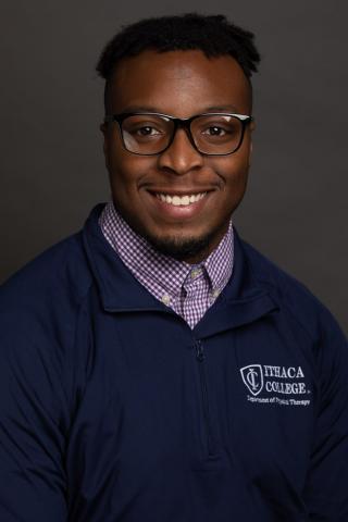 This is a photo of Olutayo Akinboboye. Olu is wearing a blue Ithaca College shirt over top of a white and blue checked shirt. Olu is wearing glasses.