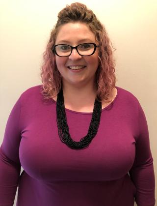 This is a photo of Sarah O'Hagen. Sarah is wearing a long sleeve purple shirt and a long black necklace around the neck. Sarah is wearing glasses.