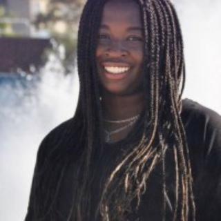 This is a photo of Temilola Adeoye. Temi is standing in front of the Ithaca College fountains wearing a long black shirt and is smiling for the camera.