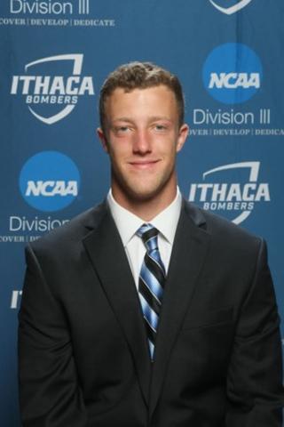 This is a photo of Tyler Winslow. Tyler is wearing a dark blue suit with a white shirt and blue striped tie. Tyler is standing in front of an Ithaca Athletics Division III background.