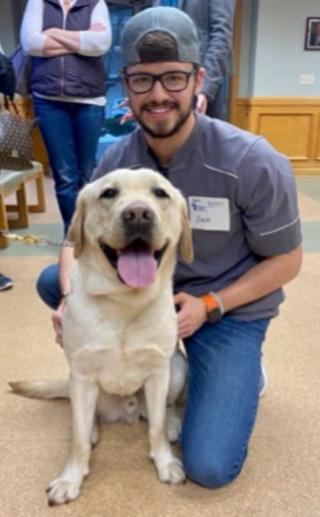 This is a photo of Zachary Thomas. Zachary is kneeling down petting a white dog. Zachary has glasses and is wearing a short sleeve blue shirt and blue jeans. Zachary is wearing a cap backwards.
