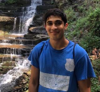 A young man stands in front of a waterfall with a blue and white shirt with a closed mouth grin