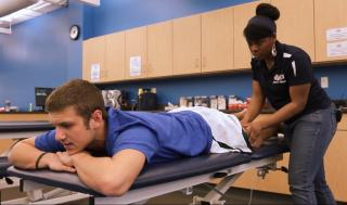 An athletic training student is standing up massaging the leg of an athlete who is laying face down on a table.