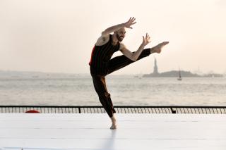 A dancer is in New York City doing a kick. The Statue of Liberty is in the background.