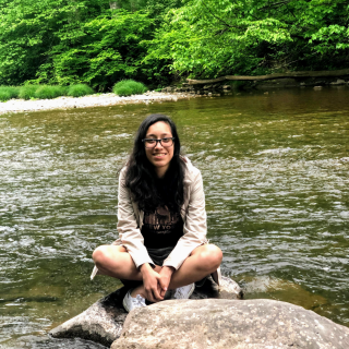 Helen is sitting on a rock in front of a body of water. She is wearing shorts and a beige hoodie, smiling at the camera.