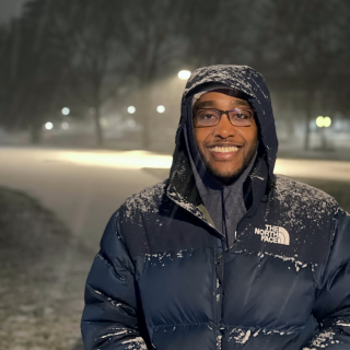 Simeon is standing in the snow in a dark blue North Face jacket with the hood pulled up. He is smiling at the camera and wearing glasses.