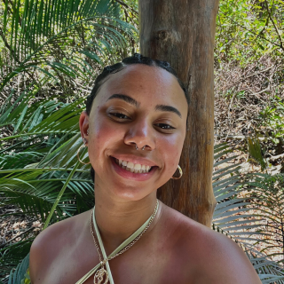 Seidy is standing in front of green plants smiling at the camera. She is wearing her hair in a series of twists, small gold hoop earrings and a criss crossed strapped blouse.