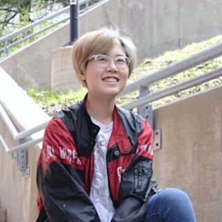 Hana is sitting on cement stairs looking up over the camera and smiling. They are wearing a read and black jacket, jeans, and a white shirt. They have on white rimmed glasses.
