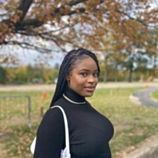 Kinza is standing outside in front of a tree looking at the camera with her body turned to the side. She is wearing a black turtleneck and her braided hair is parted to the side.