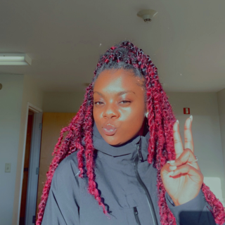 Tywanda is looking at the camera with a pursed lip and giving a peace sign with her hand. She is wearing long red twists in her hair and a grey jacket.