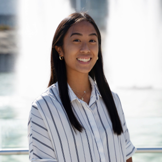 Alyssa is smiling at the camera in front of the IC Fountains. She is wearing a white and grey stripped button up shirt and her hair is parted down the middle and tucked behind her ears. 