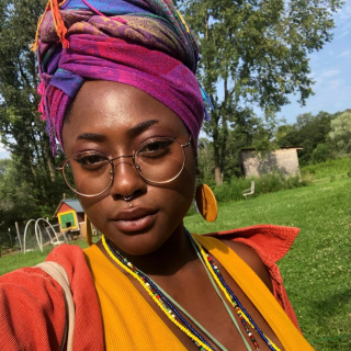 Cyepress is standing in a part. They are wearing a multicolored headwrap, orang jacket and marigold blouse. They have wooden earring and thin rimmed glasses on as they stare at the camera.