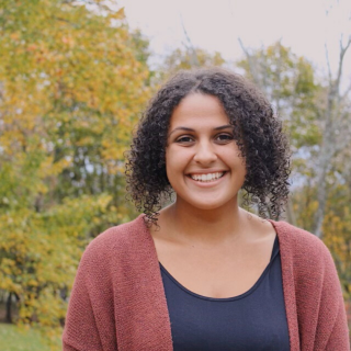 Emma is standing outside in front of autumn trees smiling at the camera. She is wearing a rust colored sweater with a navy shirt underneath. Her curly hair is parted down the middle and falls at her chin.