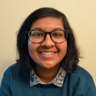 Antara is sitting and smiling at the camera. She is wearing a teal sweater with a button up collar underneath that has a blue pattern on it. She is wearing tortoiseshell glasses and her hair is parted to one side.
